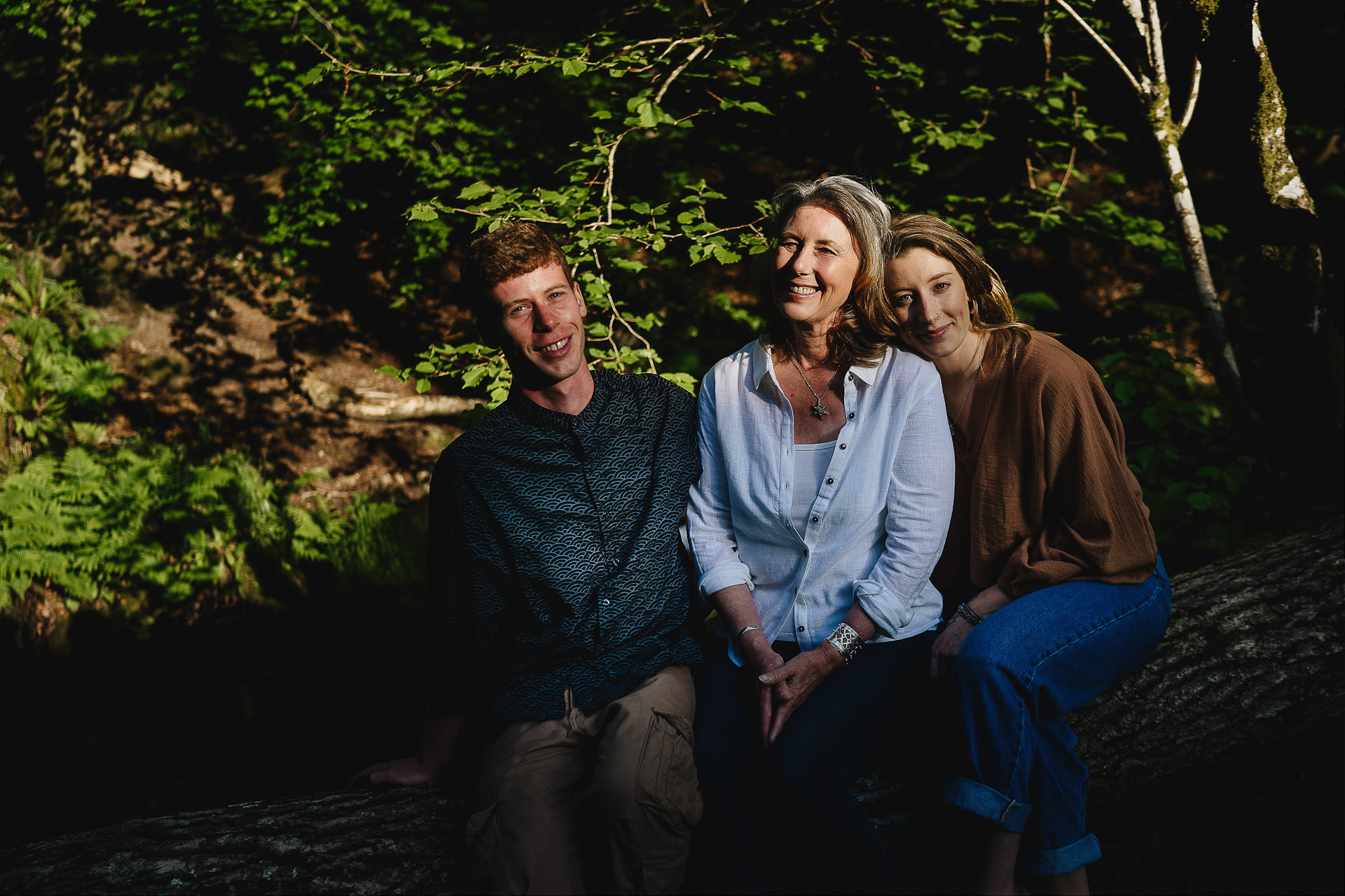 Family photography of a woman with adult children sitting on a log in woodland