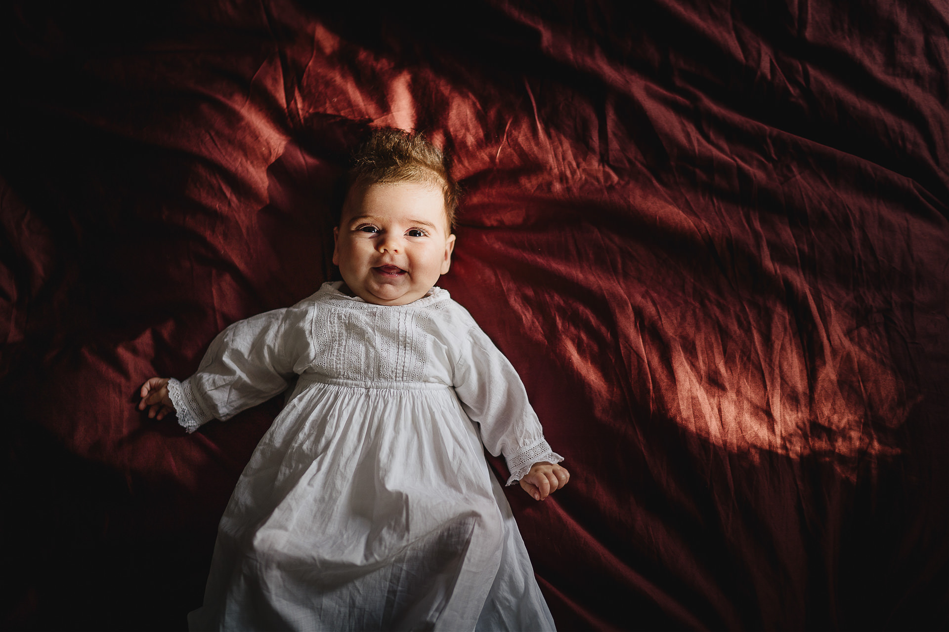 Photograph of a baby smiling on a red bed cover in the sunshine