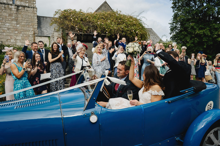 Wedding guests outside a church waving to a bride and groom in a blue vintage car