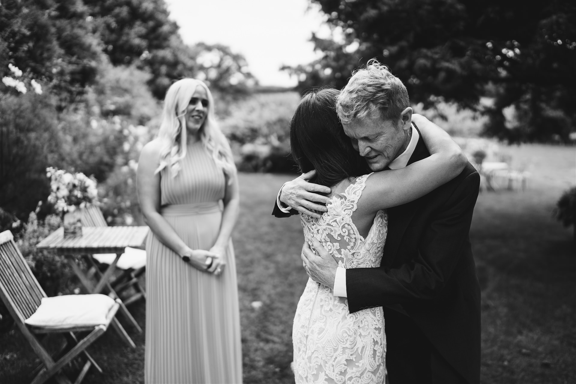 Father hugging a bride to be, with bridesmaid watching in a garden