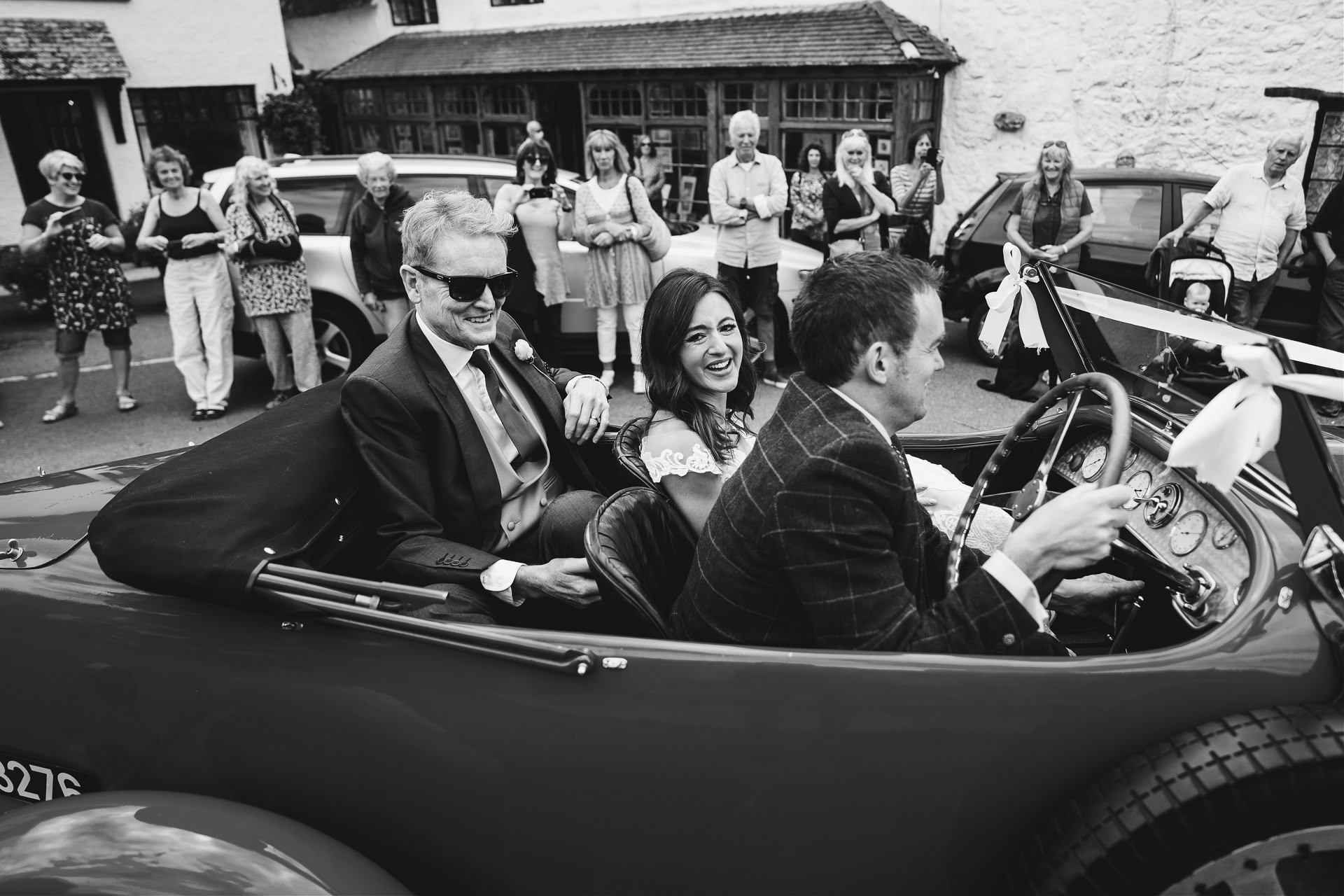 Bride and father arriving in a vintage car, with local villagers watching on the street