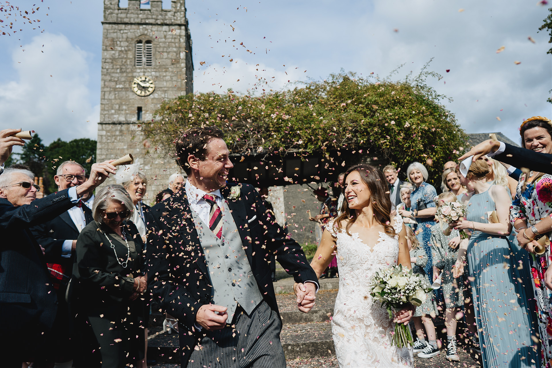 Bride and groom smiling at each other with wedding guests throwing confetti outside a church