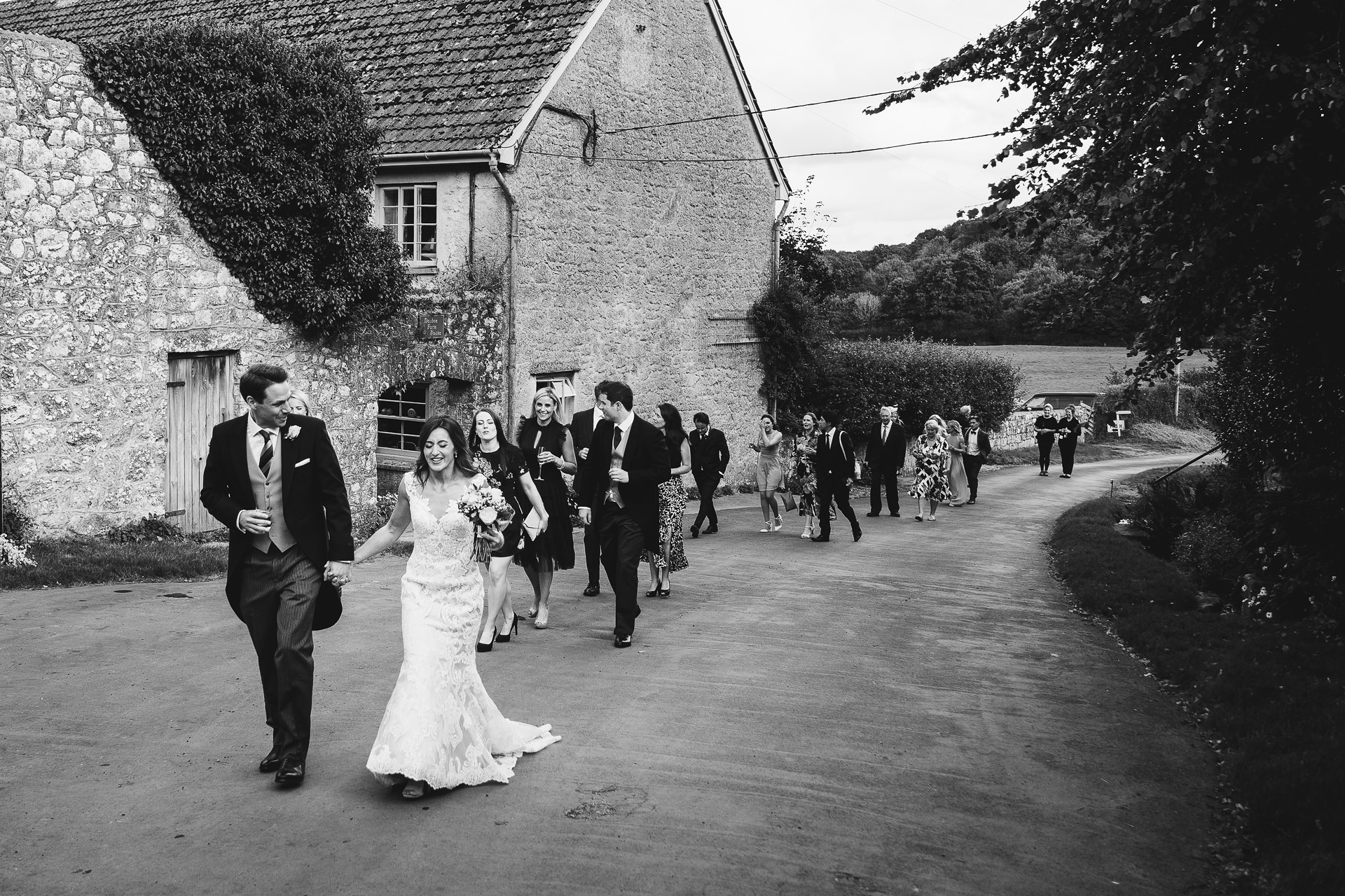 Bride and groom leading wedding guests up a beautiful country lane towards a stone barn