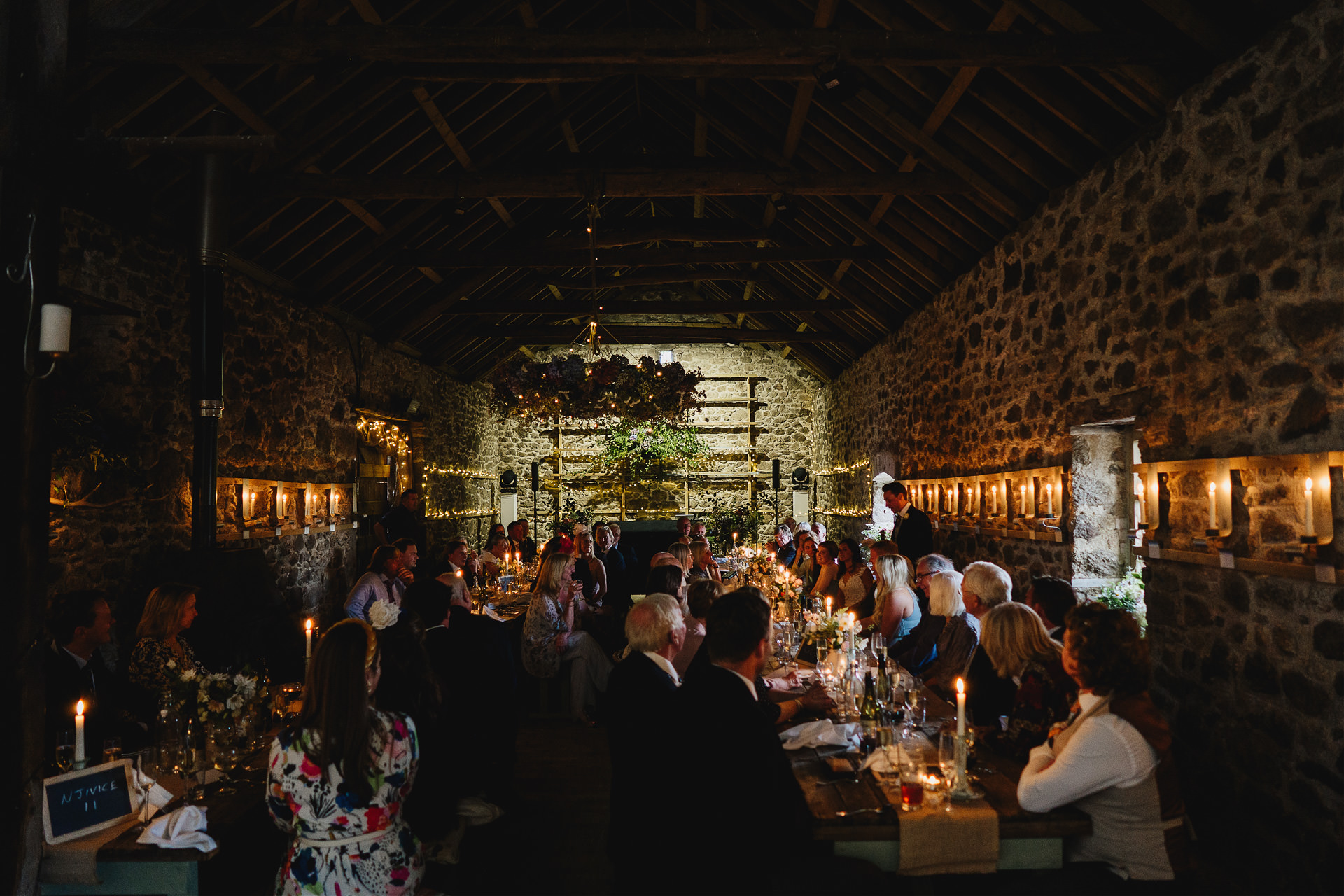 Wedding guests seated along long tables in an incredibly atmospheric wow factor wedding barn, with candles around the walls and a dark ambience