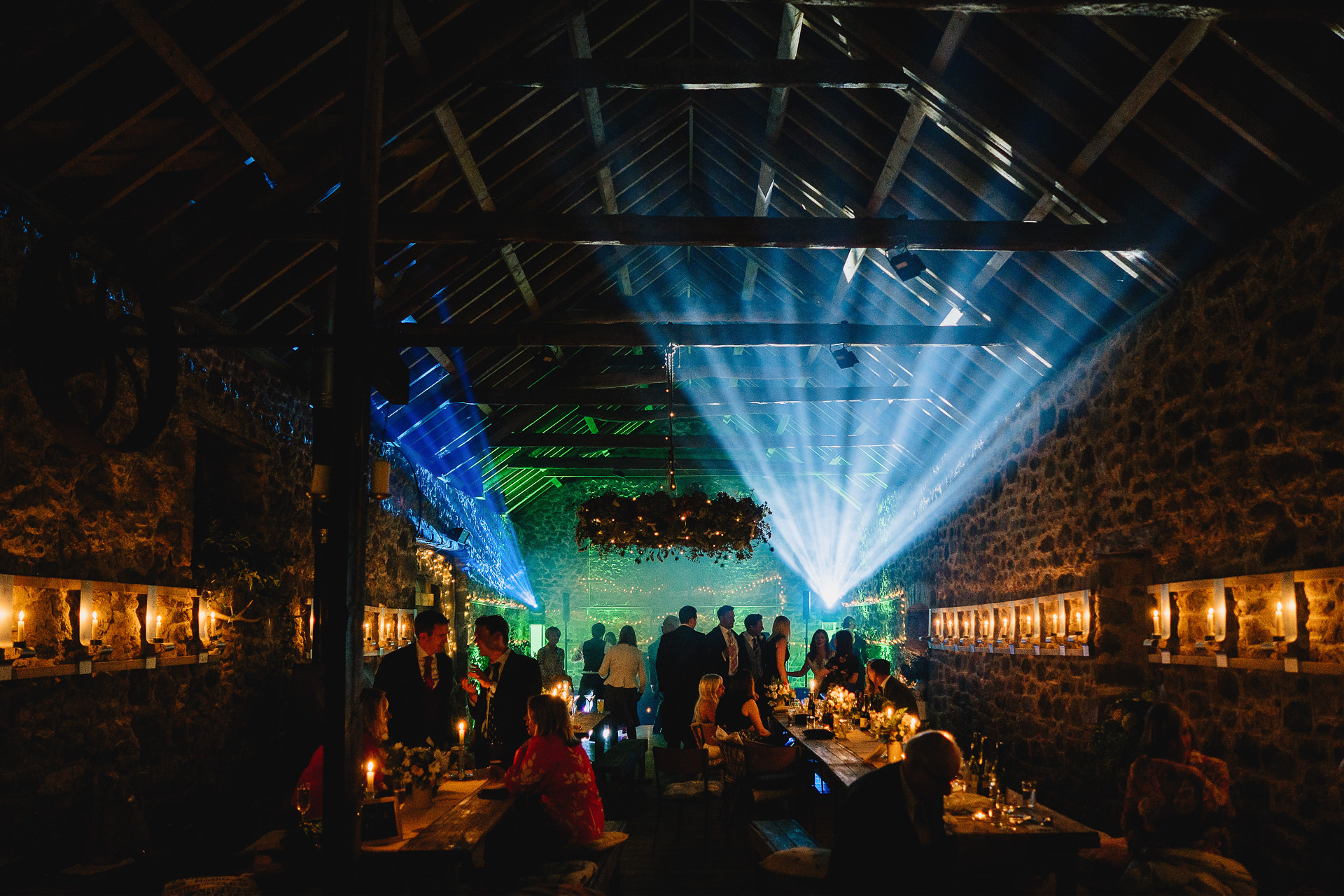 An incredible wedding barn with candles around the walls and colourful DJ lighting behind long tables 