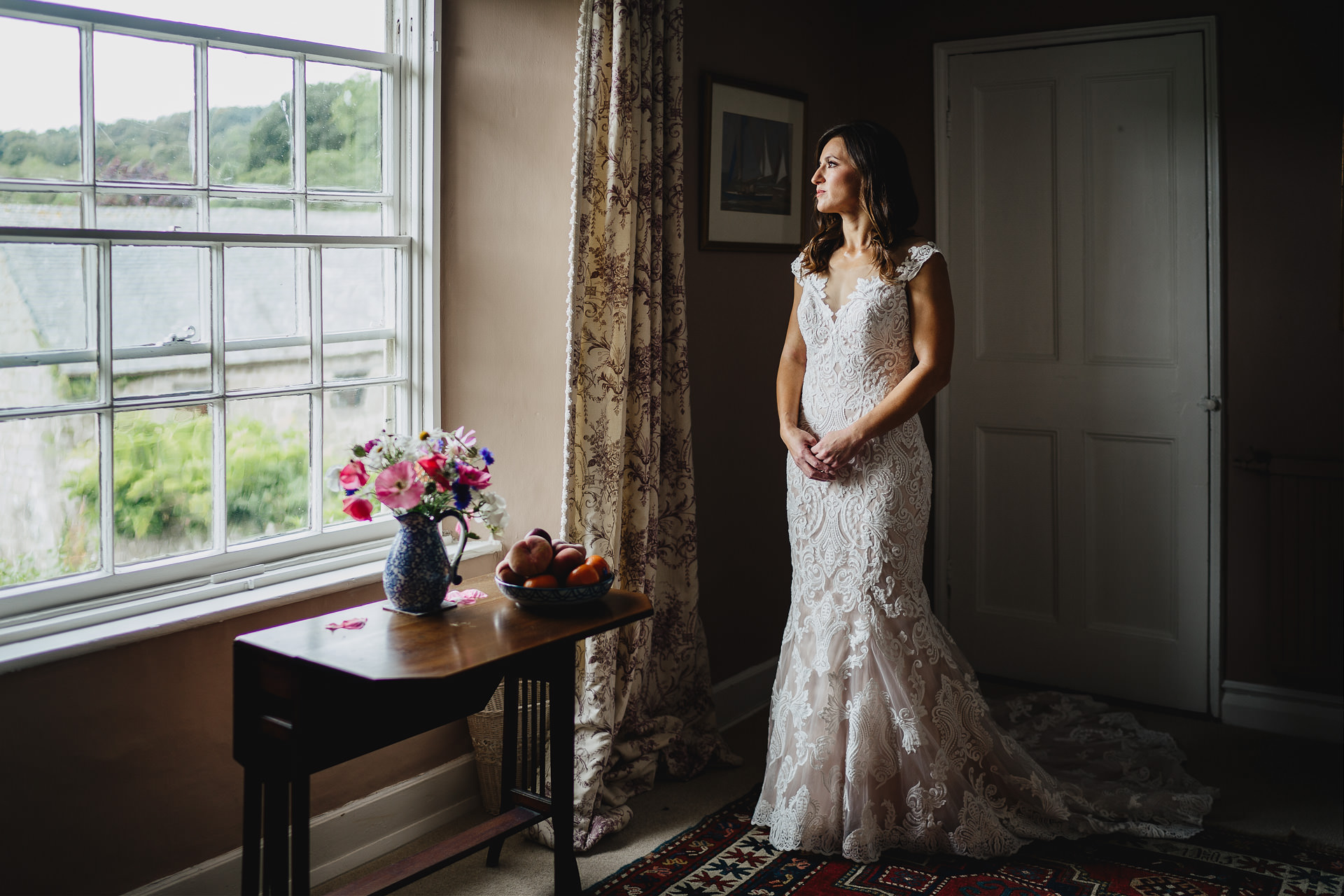 Portrait of a bride in a stunning white lace dress, standing by a window looking across Dartmoor views
