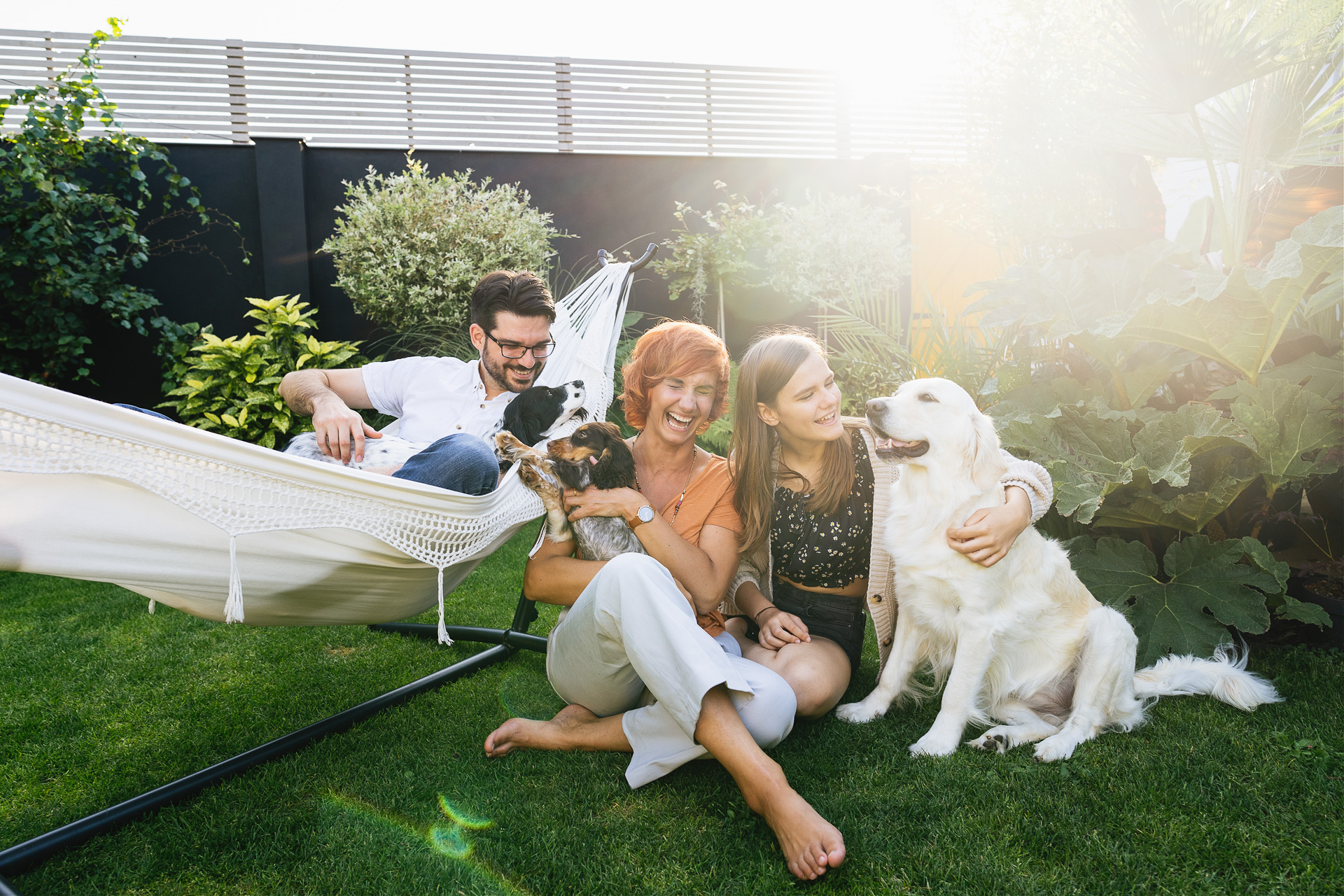A family with a man, woman and teenage girl, sitting in a garden together with three dogs. The people are al laughing. 