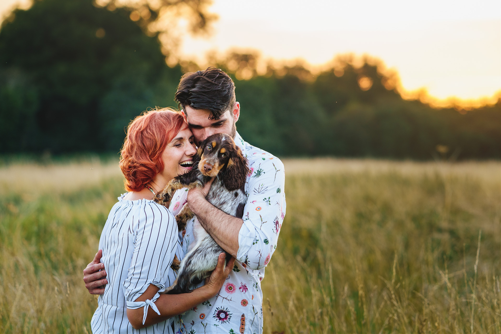 A couple cuddling with a small spaniel puppy in a field at sunset. 