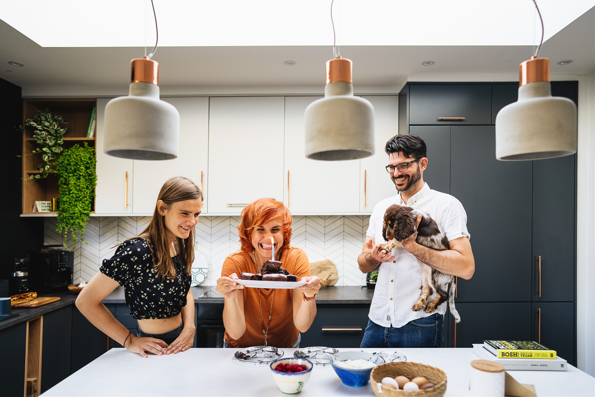 A woman laughing with a candle in a cake, with a man and dog watching and teenage girl, all standing in a beautiful kitchen together.