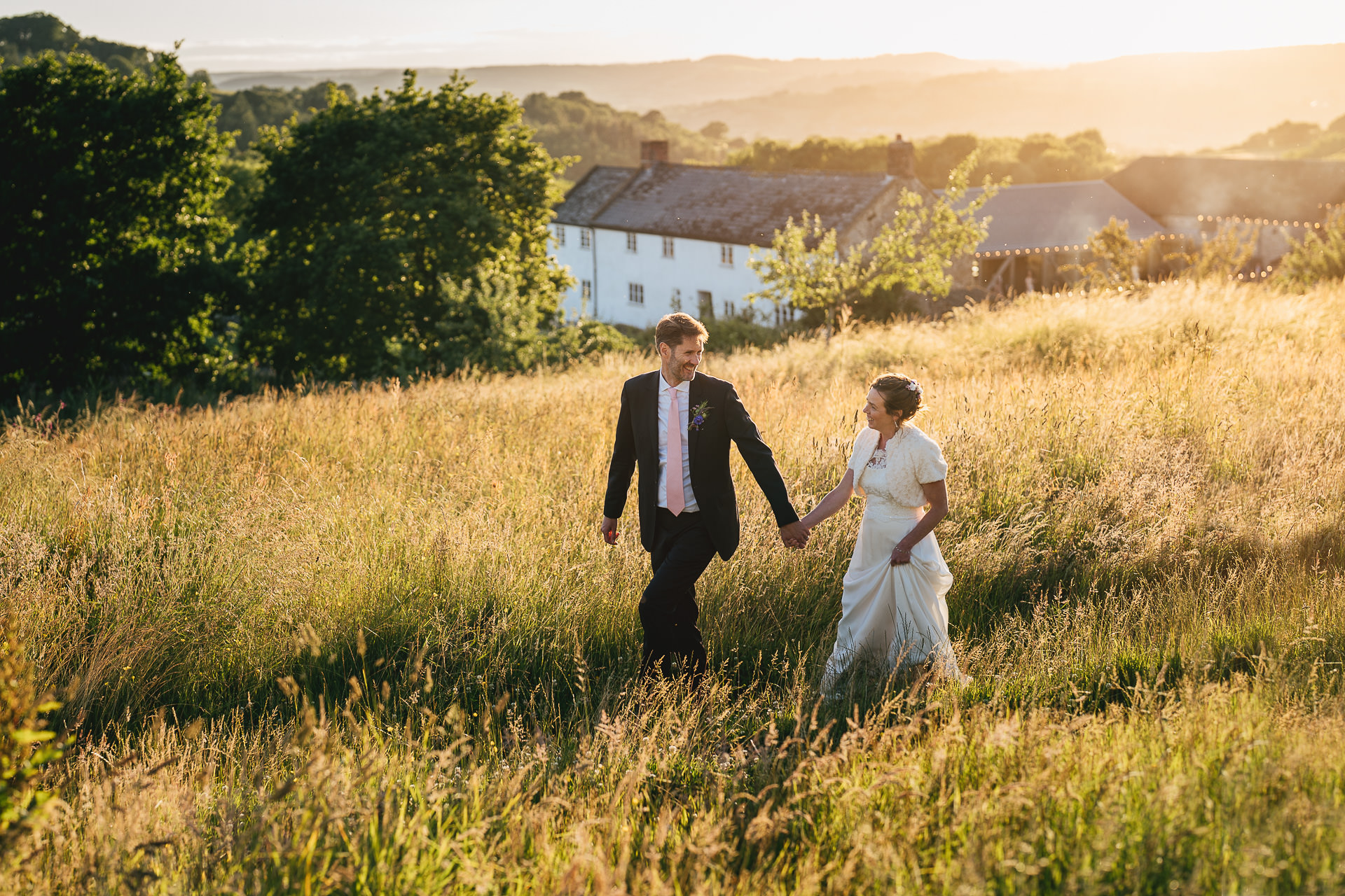 A bride and groom walking hand in hand through a grassy field at their River Cottage wedding with the farmhouse behind them and golden setting sunlight on the hills