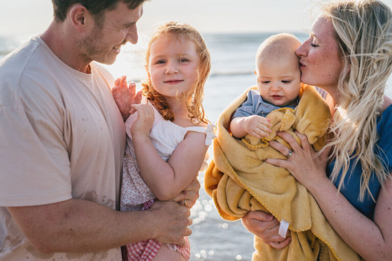 A mother and father standing on a beach cuddling two children, with the sea behind them and golden sunlight