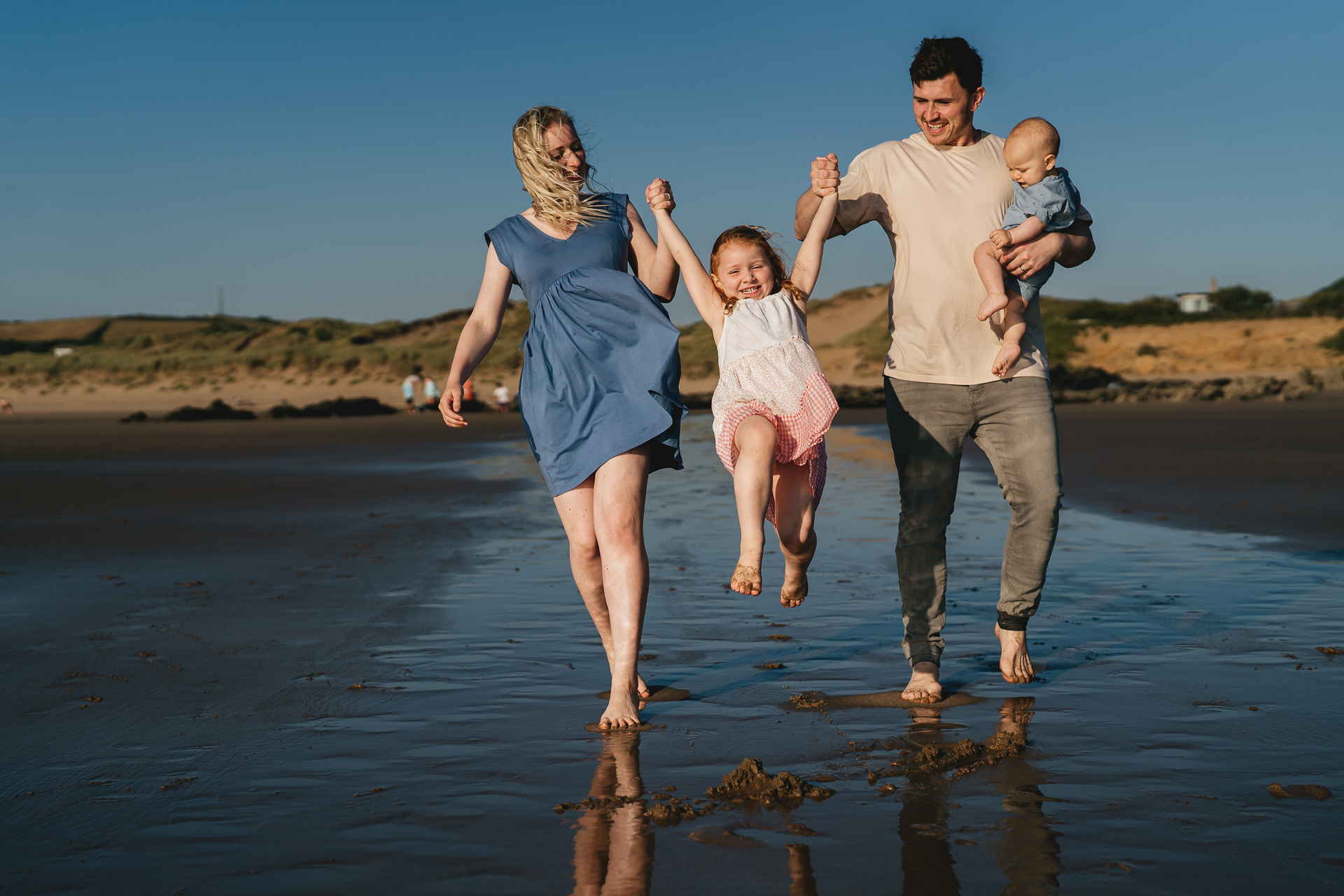 A family having fun together on the beach in North Devon, with two parents swinging a young girl between them whilst carrying a baby