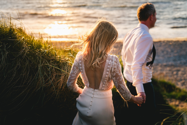 A groom and bride walking through sand dunes towards a beach with the sun setting over the sea. Bride is wearing a backless Laure de Sagazan wedding dress. 