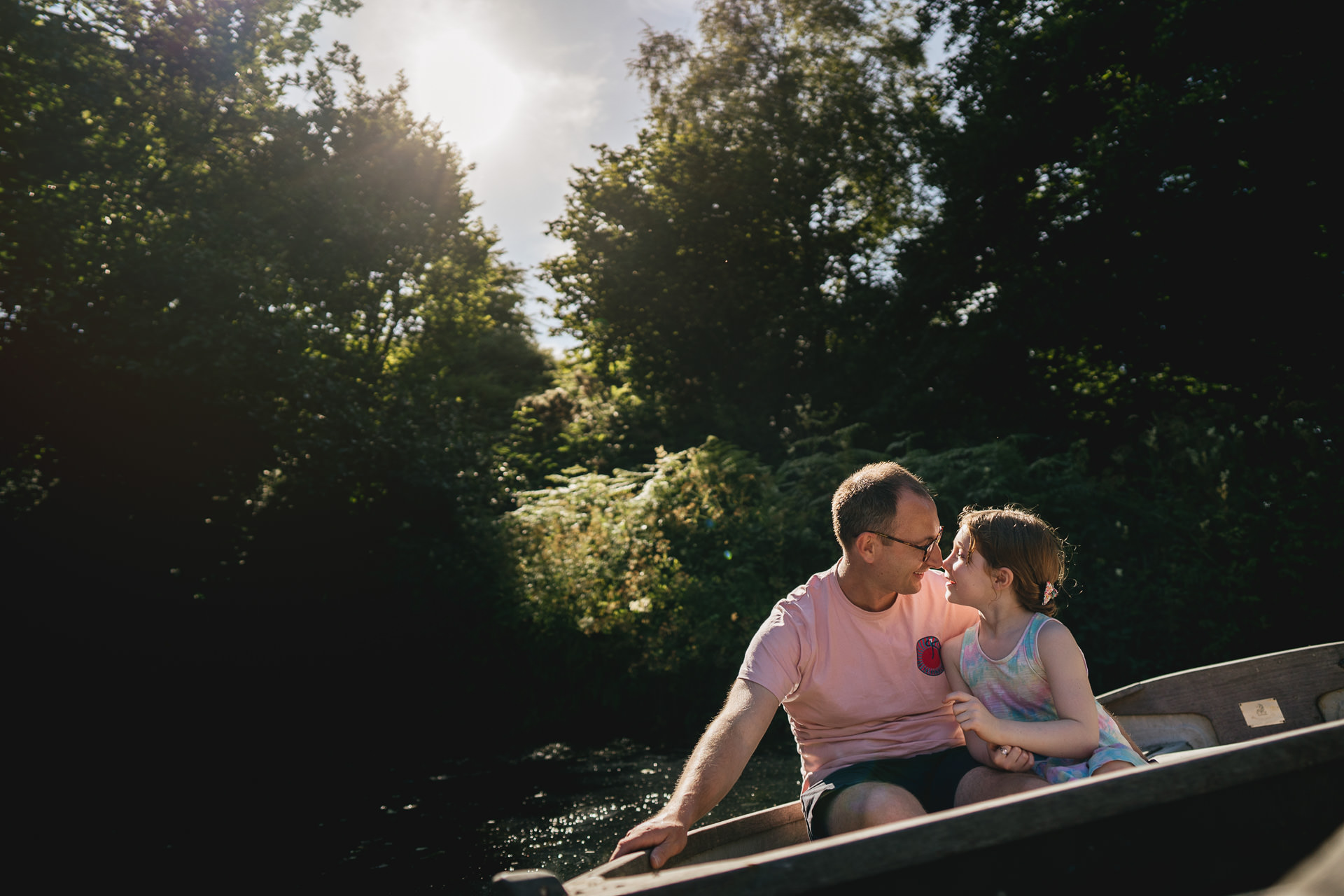 A father and daughter sitting in a small boat together in the sunshine, smiling at each other and touching noses