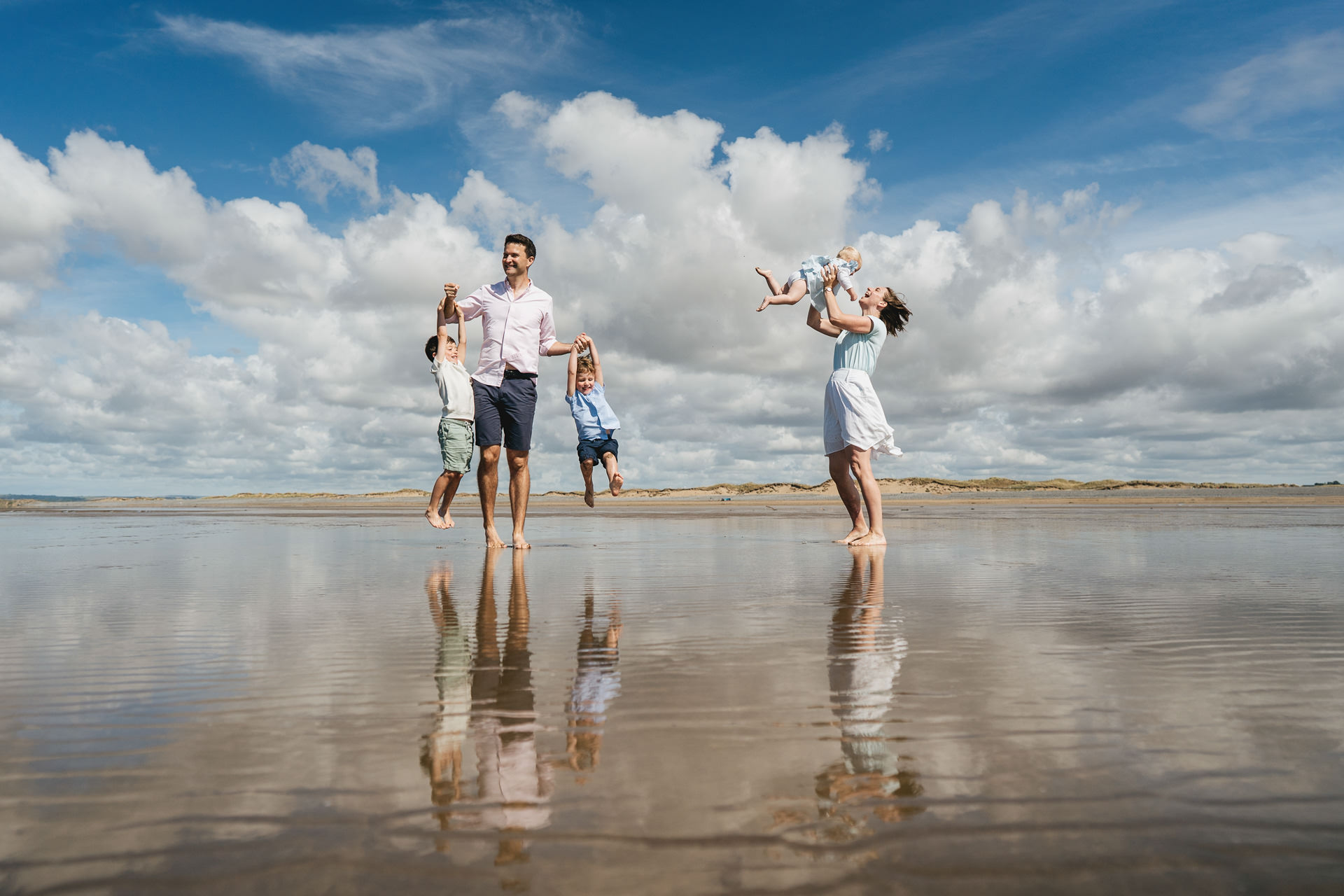 A mother and father with three young children, swinging them in the air on a beach, with blue skies above reflected in wet sand