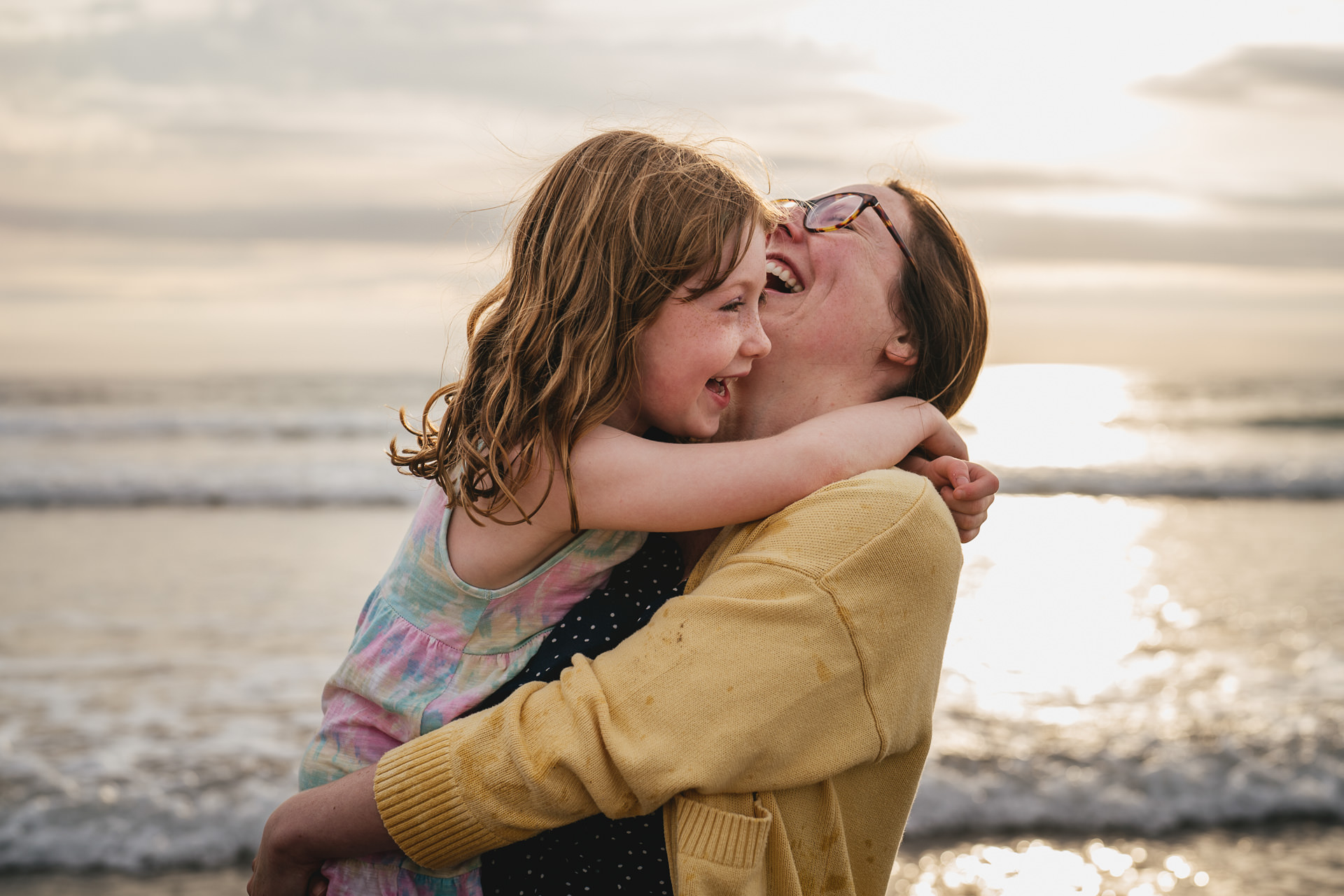 A mother and daughter cuddling and laughing together with the sun setting over the sea behind them