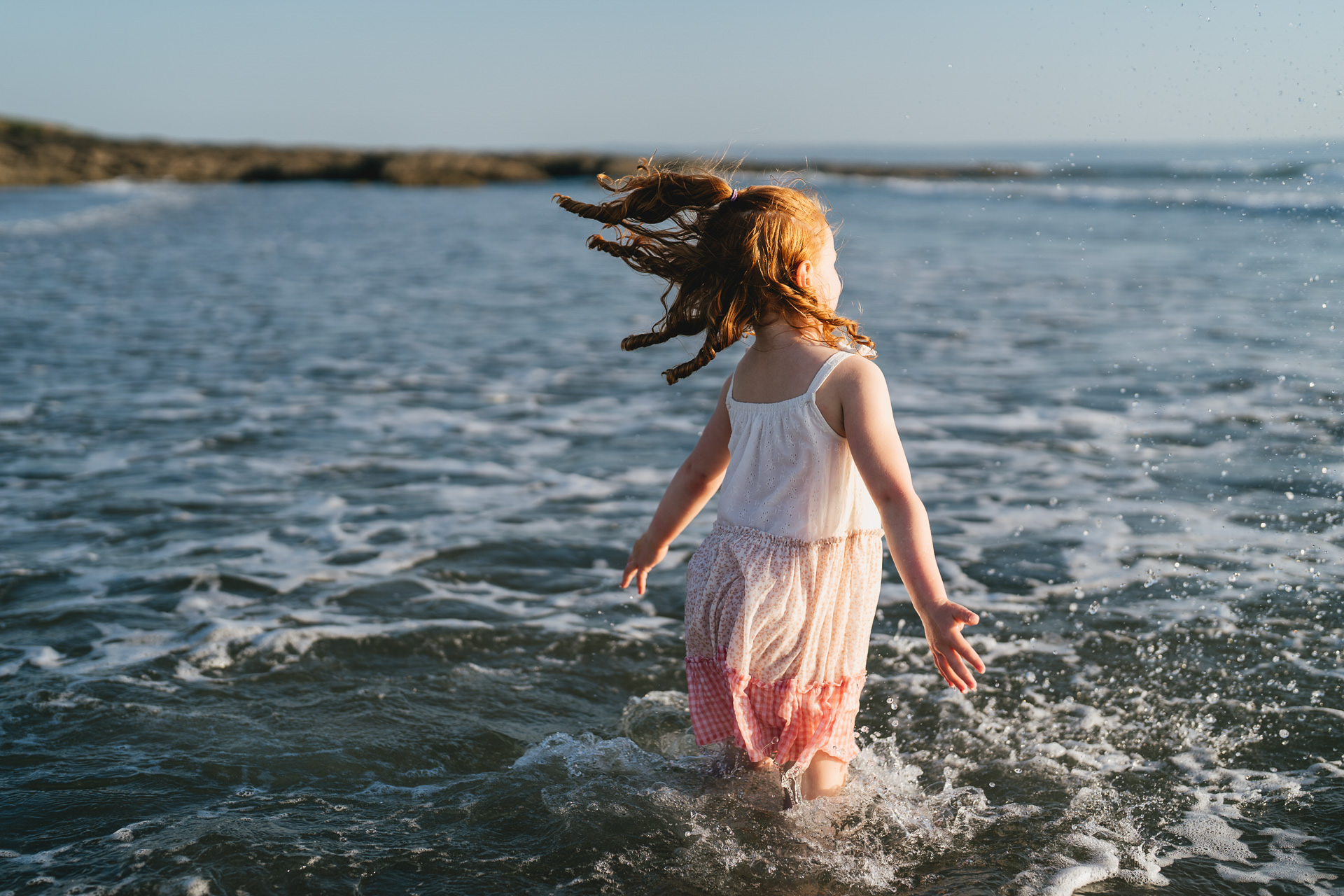 A young girl with ginger curls and a pink dress, jumping through waves in the sea