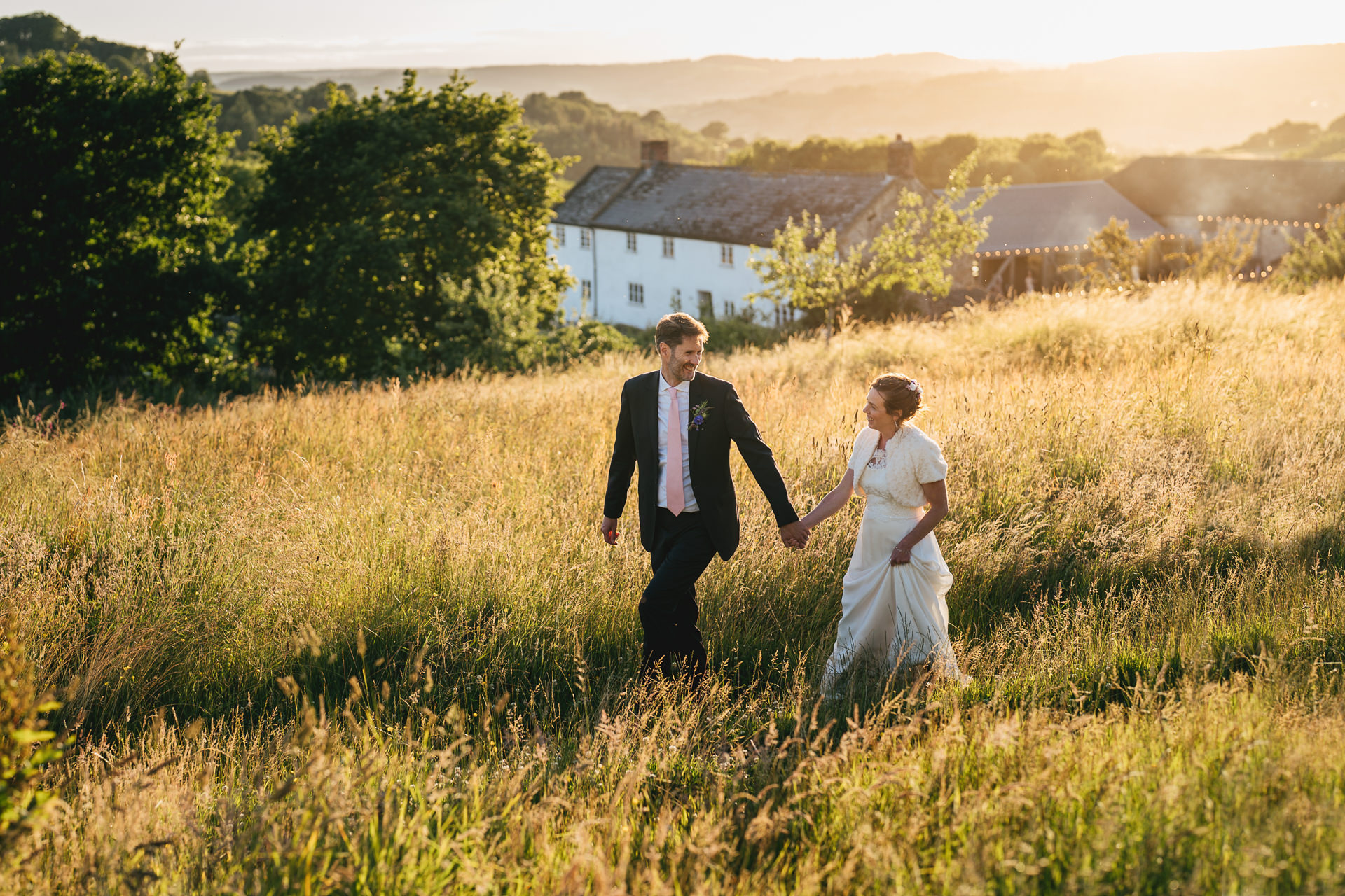 A groom and bride walking together through long grass with River Cottage behind them and the sun setting over the valley