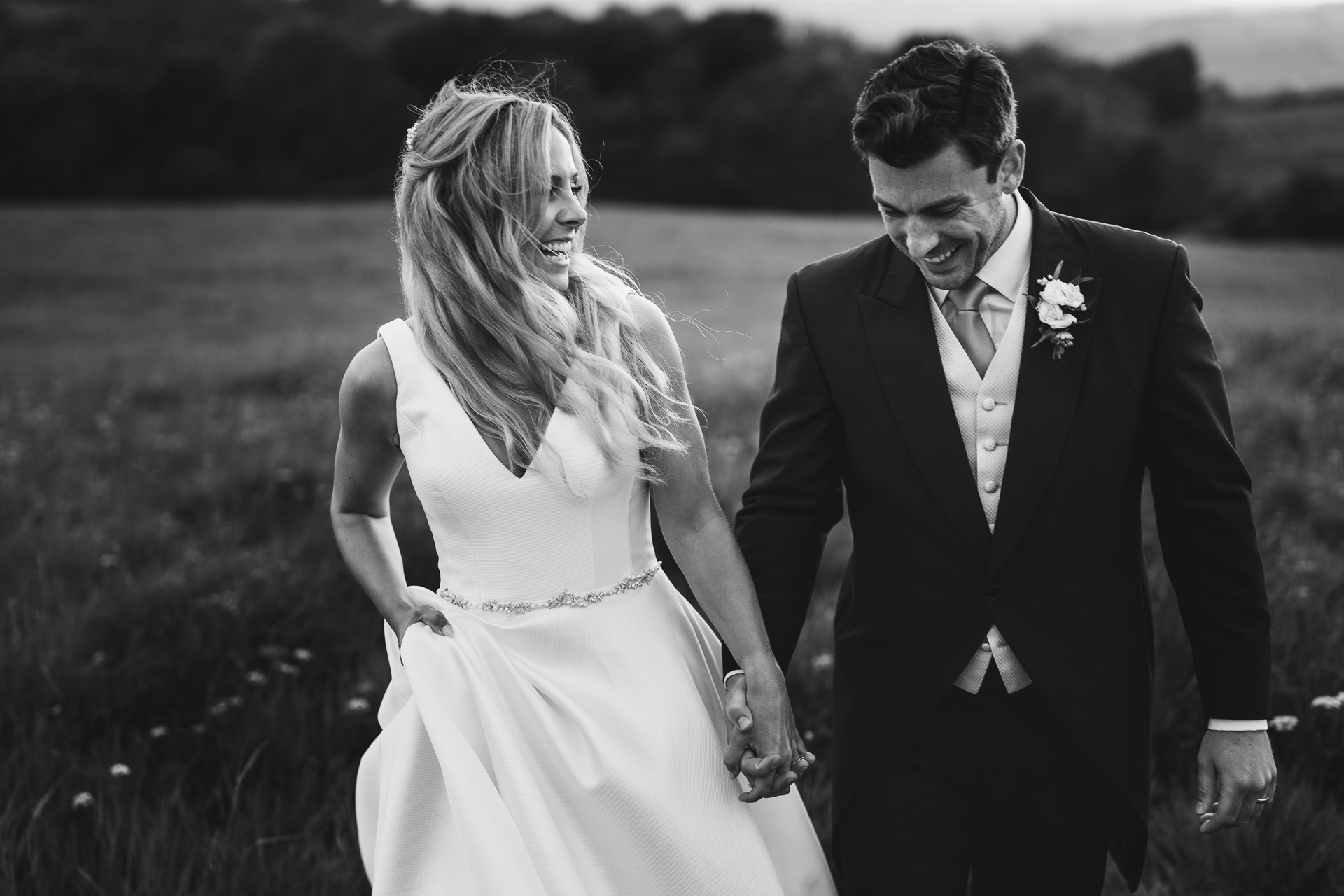 Black & white image of a bride and groom walking hand in hand across a field and smiling at each other
