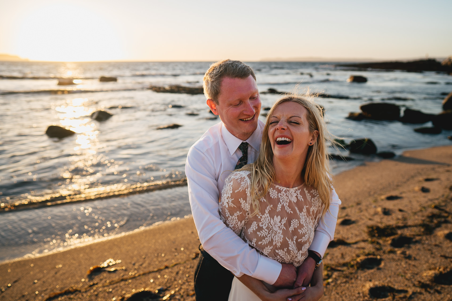 A bride and groom cuddling and laughing together on a beach at sunset