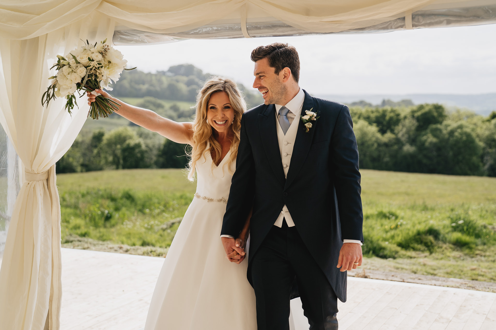 A smiling bride and groom entering a marquee and waving a bouquet, with fields, trees and hills behind them