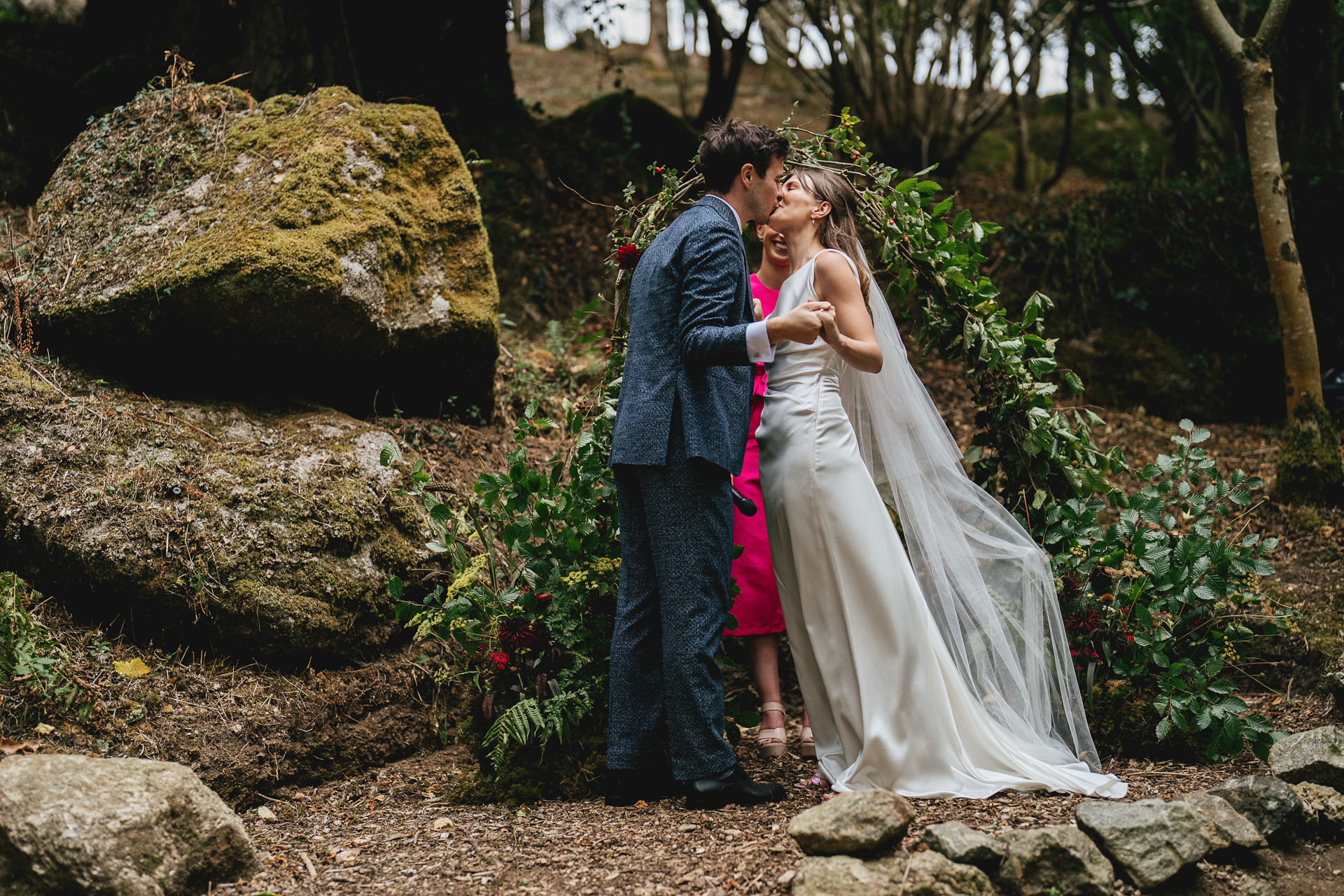 A bride and groom just married in an outdoor woodland ceremony, kissing each other by a hazel arch