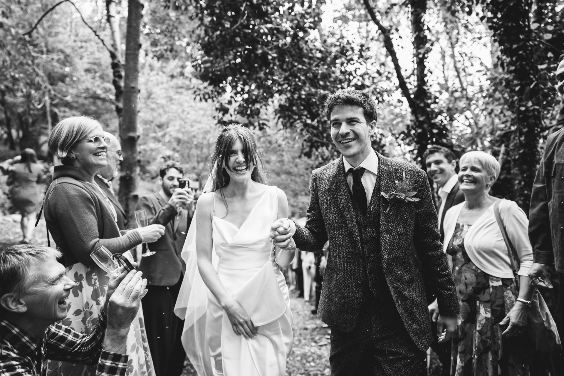 A bride in Vivienne Westwood and groom in a bespoke Casely Hayford suit laughing as guests throw confetti at them in the woodland on Dartmoor