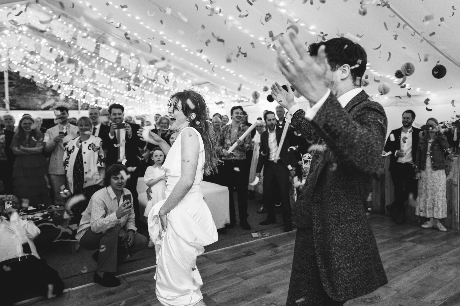 A bride and groom laughing with a confetti canon exploding above them and wedding guests watching