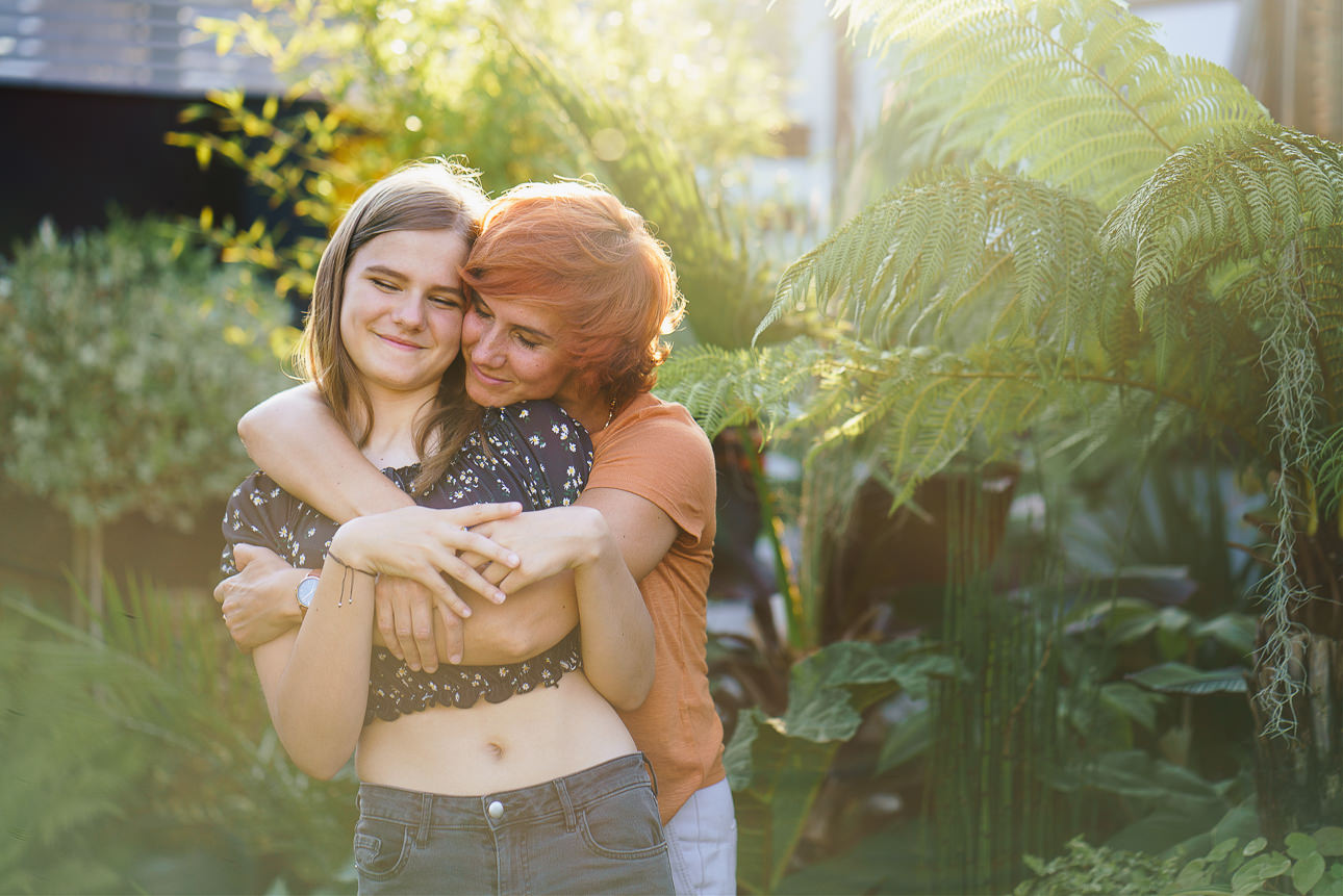 A mother and teenage daughter cuddling in soft sunlight with green ferns behind them