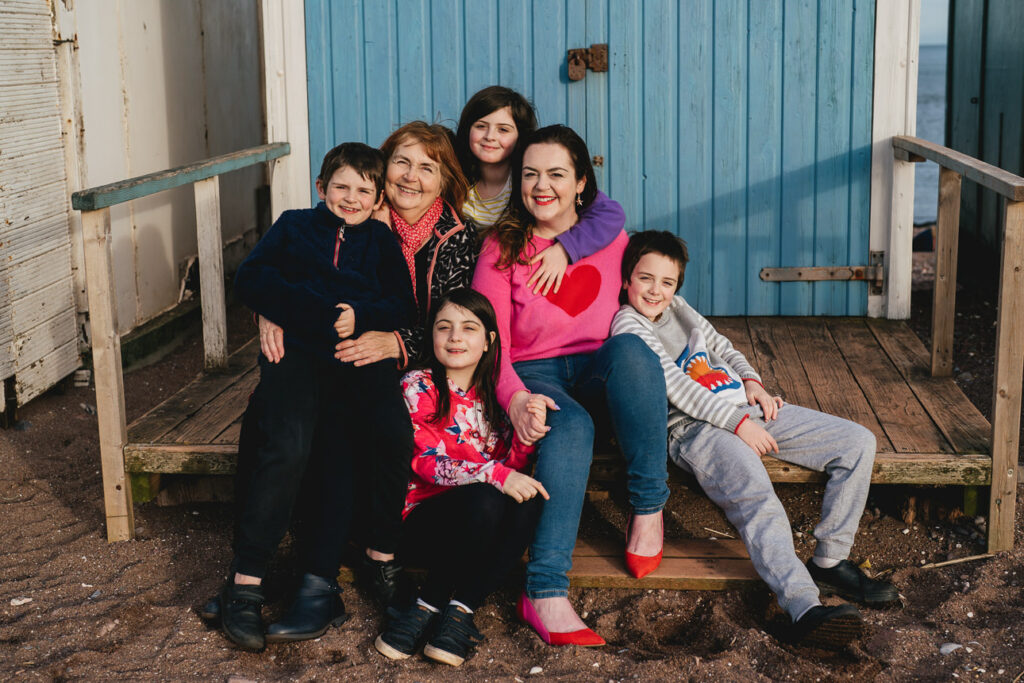 A grandmother, mother and four children smiling together in front of a blue beach hut
