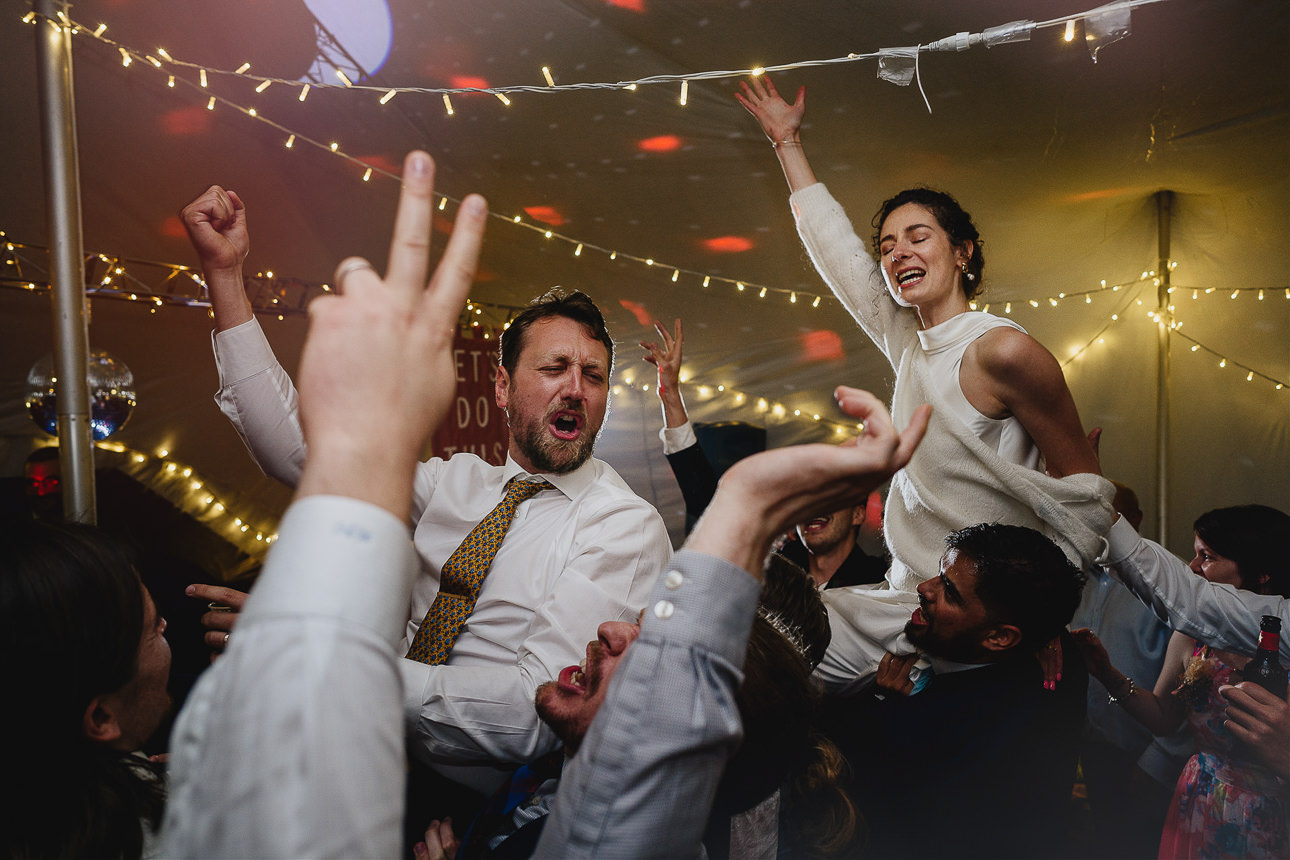 A stylish bride and groom being carried by wedding guests on the best wedding dance floor that ever existed