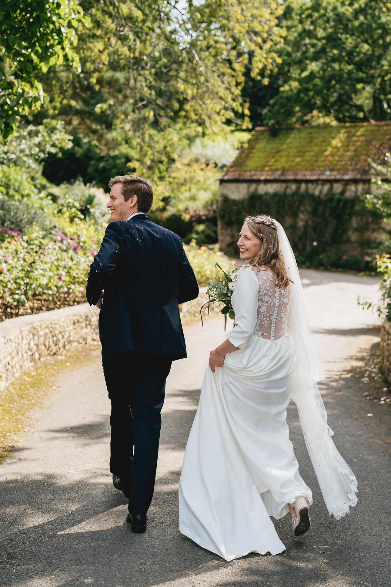 A bride and groom smiling and walking down a country lane together in sunshine after their Devon wedding ceremony