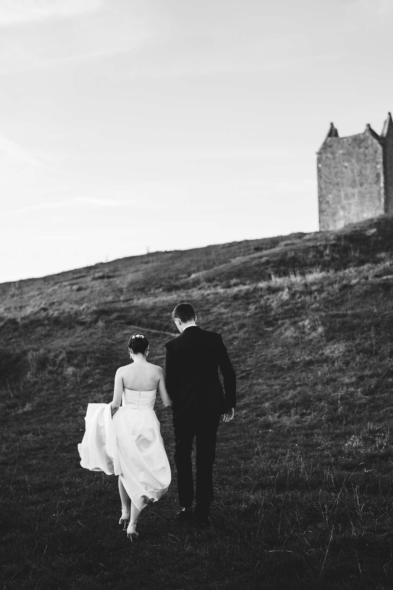 Relaxed black & white photo of a bride and groom walking up a hill together hand in hand with the Bruton dovecote above them