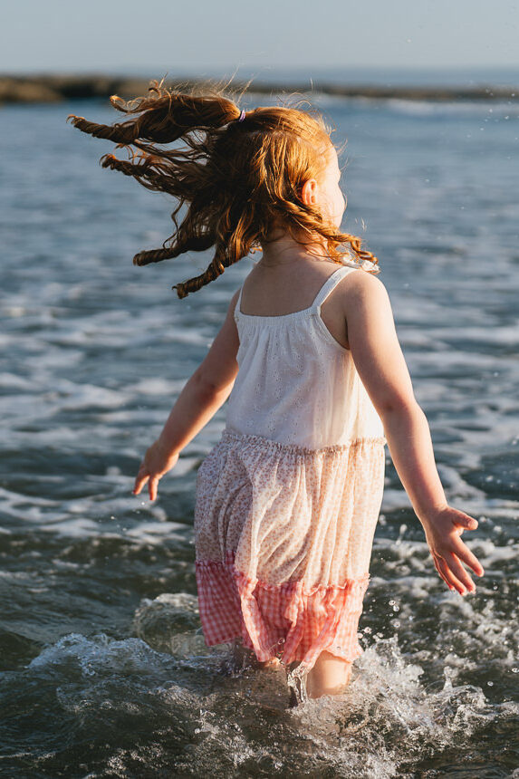 A young girl jumping in the sea with red hair and curls flying around her head