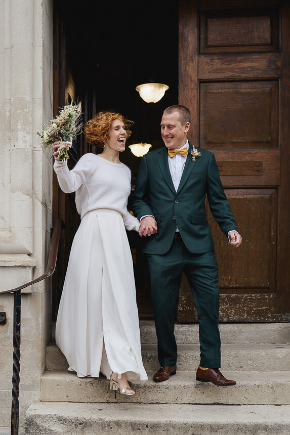 A stylish bride and groom leaving the register office after their elopement wedding at Cardiff City Hall. They are smiling and the bride is waving her bouquet in the air.
