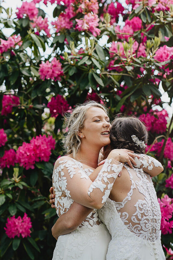 Two brides cuddling together in front of colourful rhododendron bushes