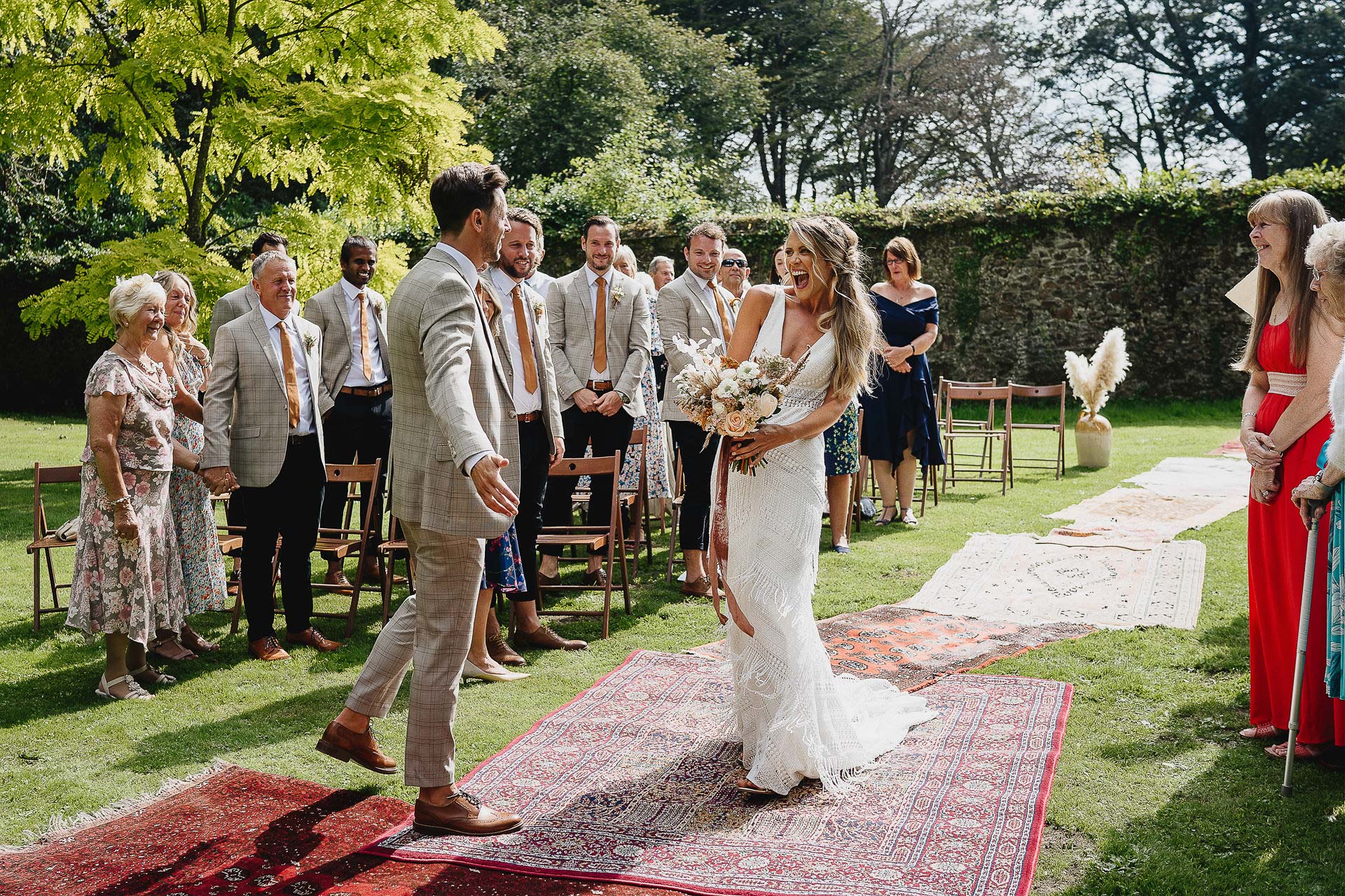 A boho bride and groom smiling at each other at their outdoor wedding ceremony in a walled garden in Devon