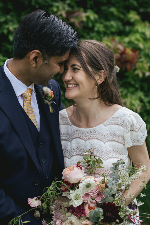 A bride and groom cuddling together in front of a leafy bush. Groom in a stylish bespoke suit and bride in a beautiful Laure de Sagazan dress
