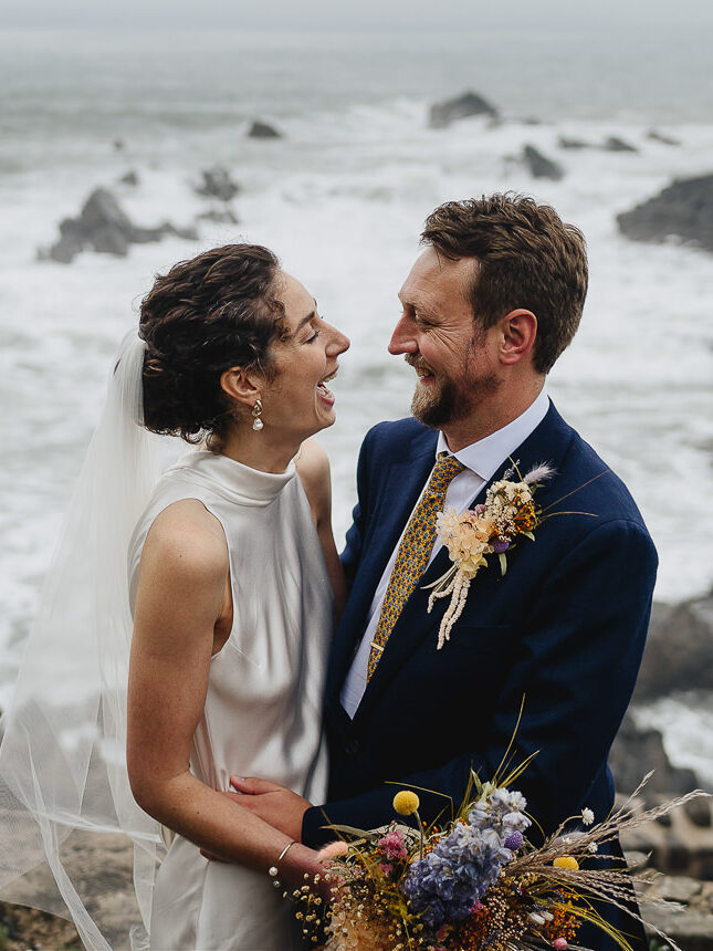 A bride and groom cuddling together in front of the Devon sea and laughing