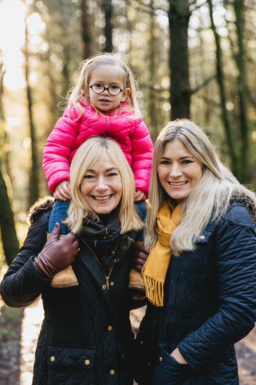 An extended family group photo, with mother, daughter and granddaughter smiling together in the woodland