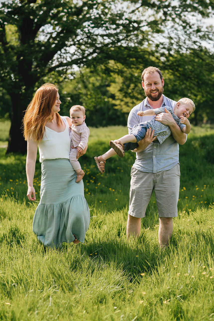 Two parents walking across grass with two young children in their arms during a family photography session in Exeter, Devon