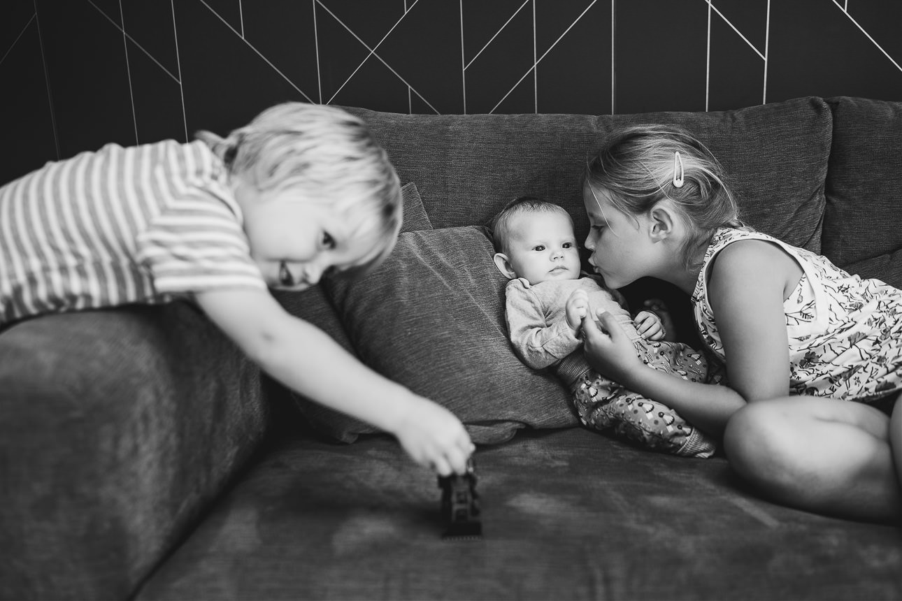 A baby sitting on a sofa with older siblings. The sister is kissing the baby and the younger brother is playing with a car. 