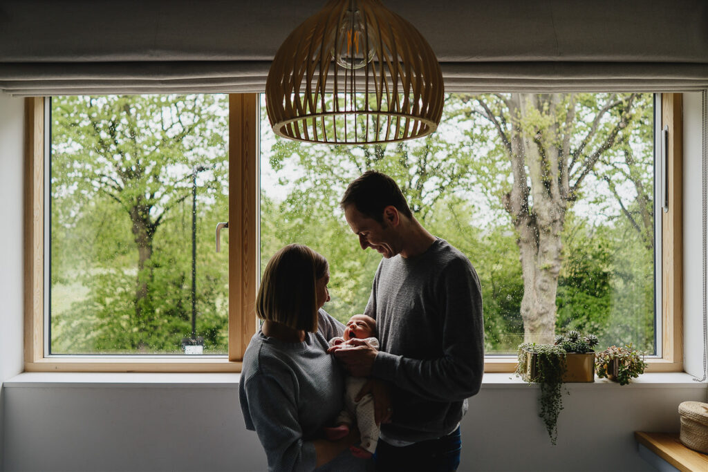 A mother and father holding a newborn baby in front of a large window with trees outside
