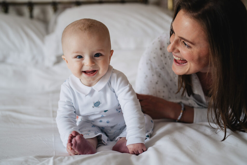 A smiling baby sitting on a bed with his mother smiling at him