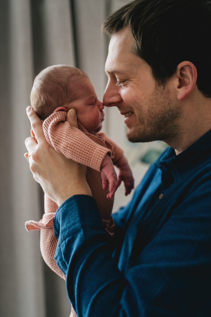 A father holding a newborn baby up to his face and smiling as they touch noses