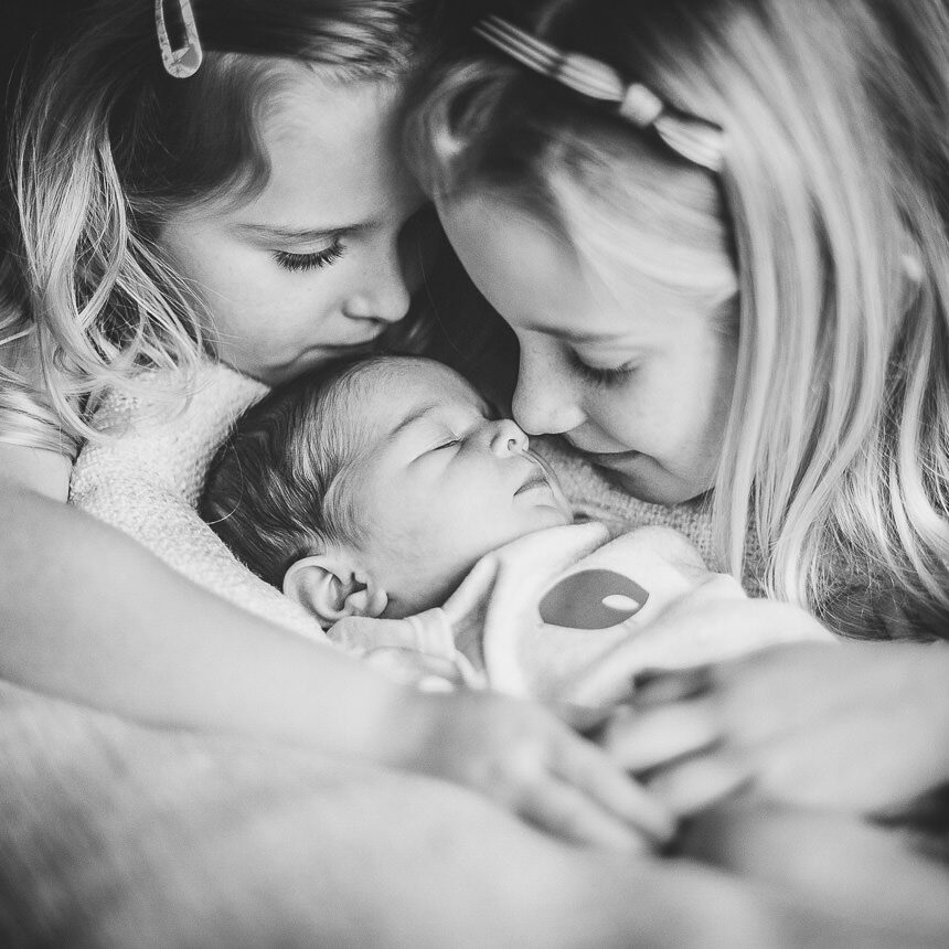 A newborn baby with two older sisters cuddling around her. One of the sisters is touching her nose to the baby. 