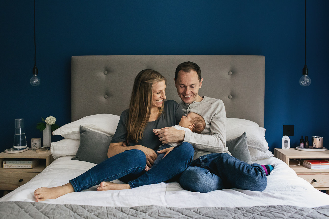 Two parents with a small baby, sitting on a double bed together in a stylish bedroom with blue walls
