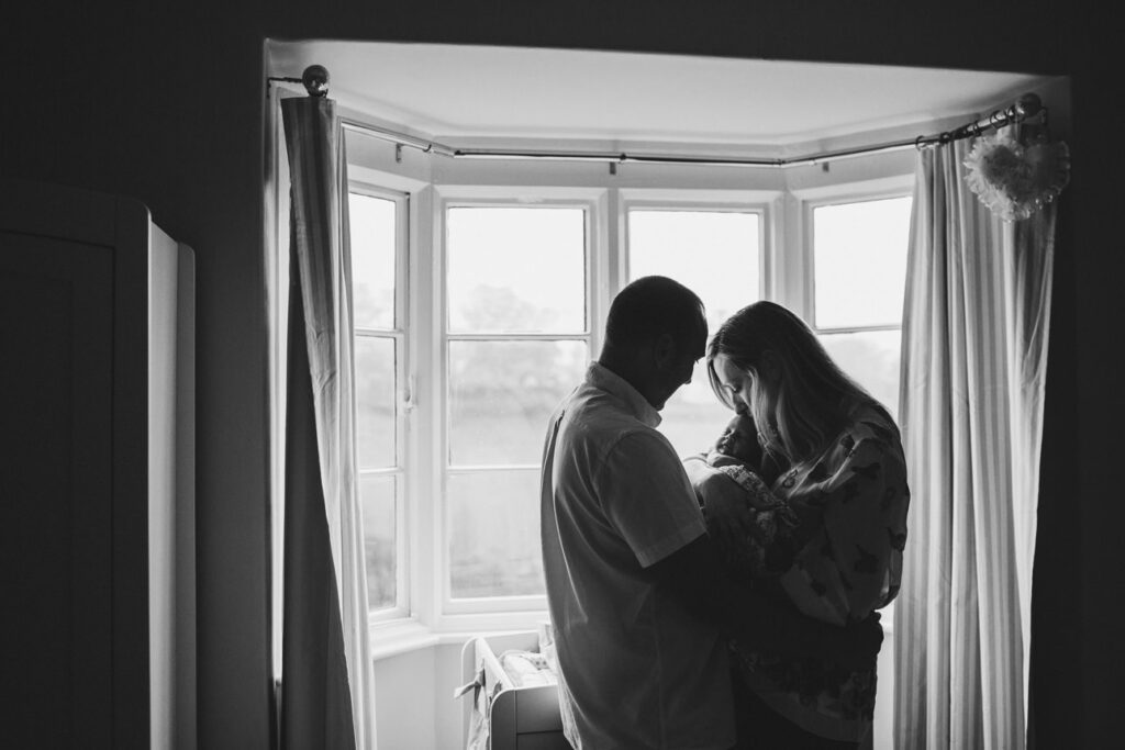 A mother and father cuddling their newborn baby in front of a large window