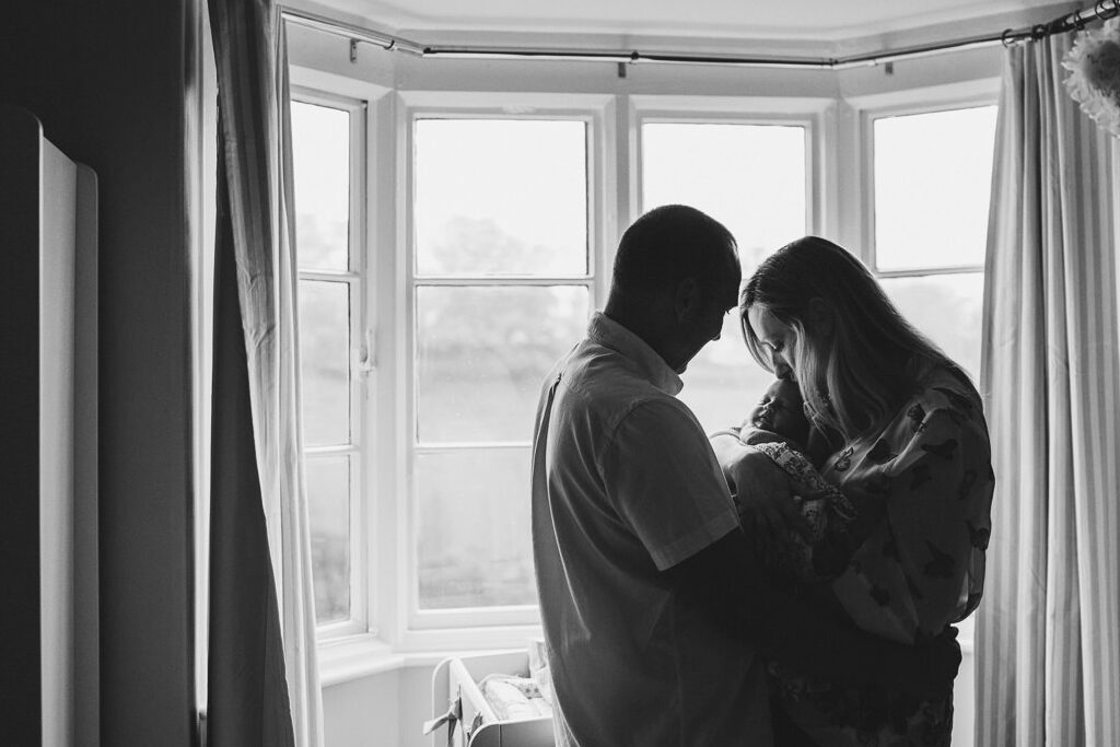 A mother and father cuddling their newborn baby in front of a large window