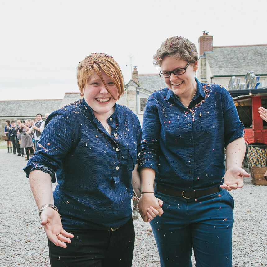 Brides in stylish blue shirts smiling as they walk through guests throwing confetti over them